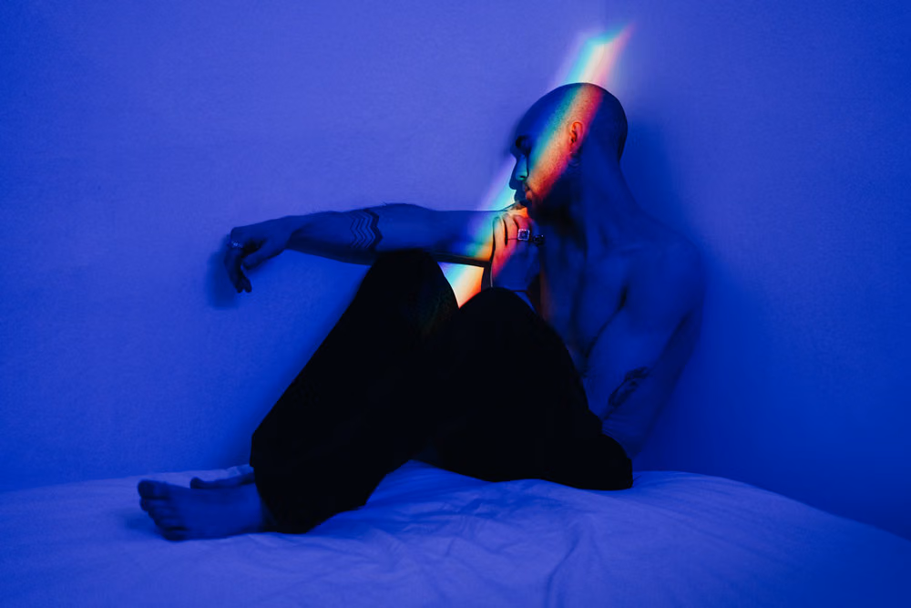 Shirtless man sitting on a bed in blue light, a streak of rainbow light across his face