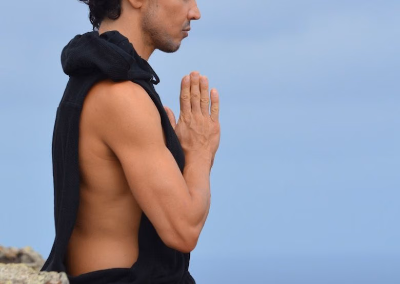 Man meditating outdoors with palms pressed together against a pale blue sky