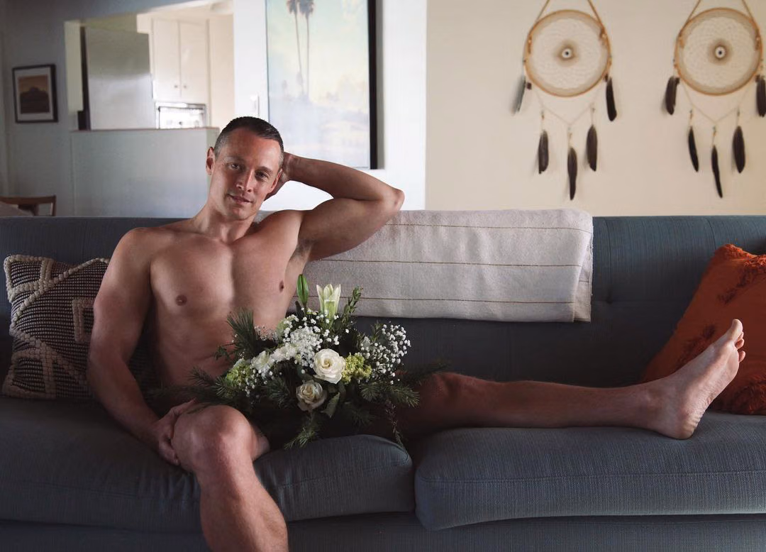 Davey Wavey posing nude on a gray sofa, one arm behind his head and a bouquet of white flowers covering his lap