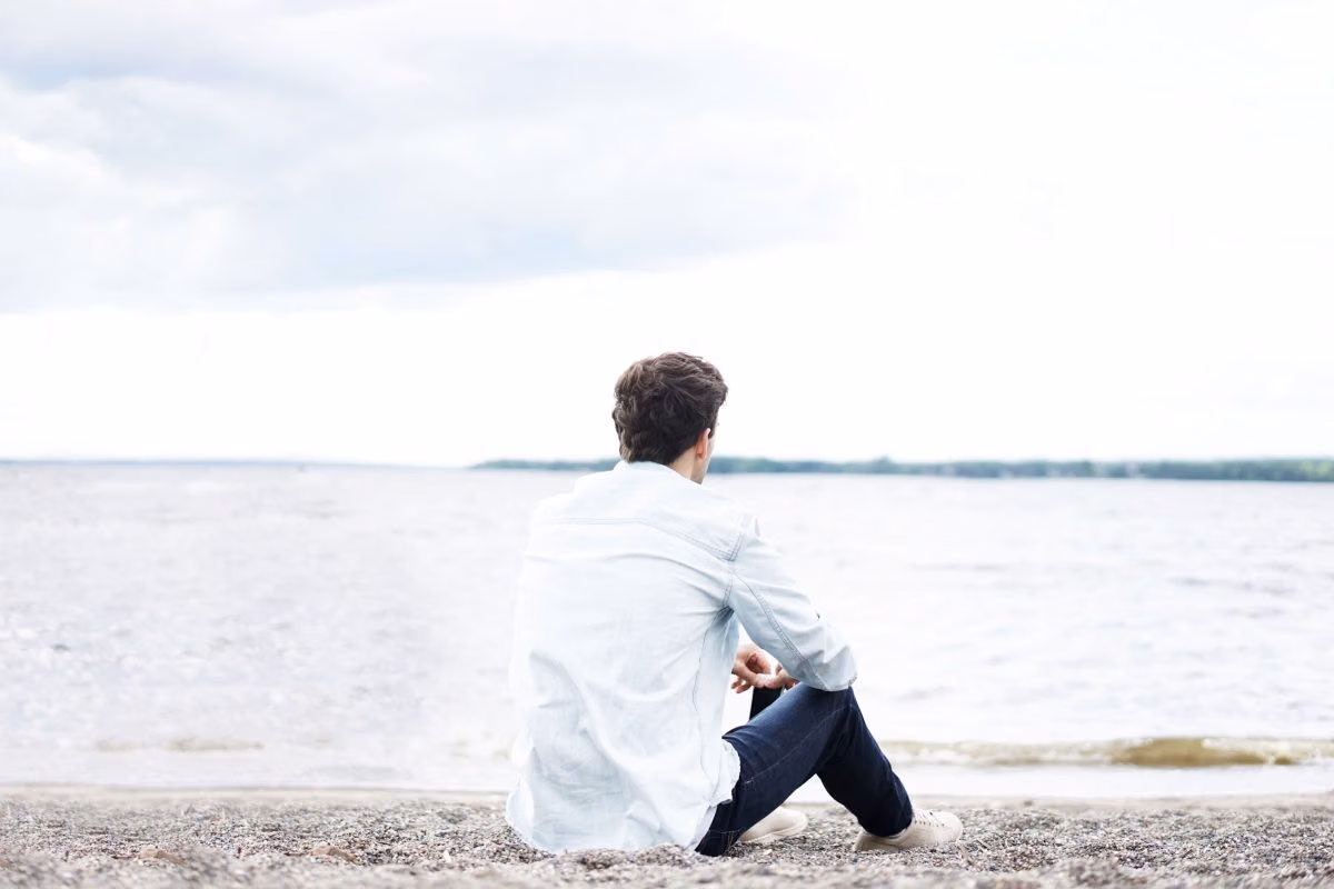 Man in a denim shirt sitting alone on a pebbly shore, looking out over gray water