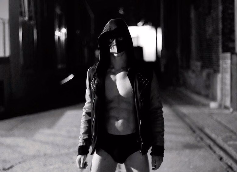 Black-and-white shot of Black Spark, masked and hooded in a studded leather jacket and black briefs, in a dark alley