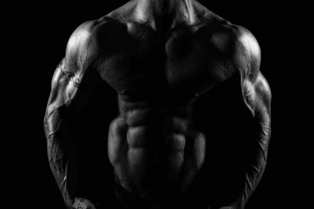 Black-and-white close-up of a muscular man's chest, shoulders and abs against a black background