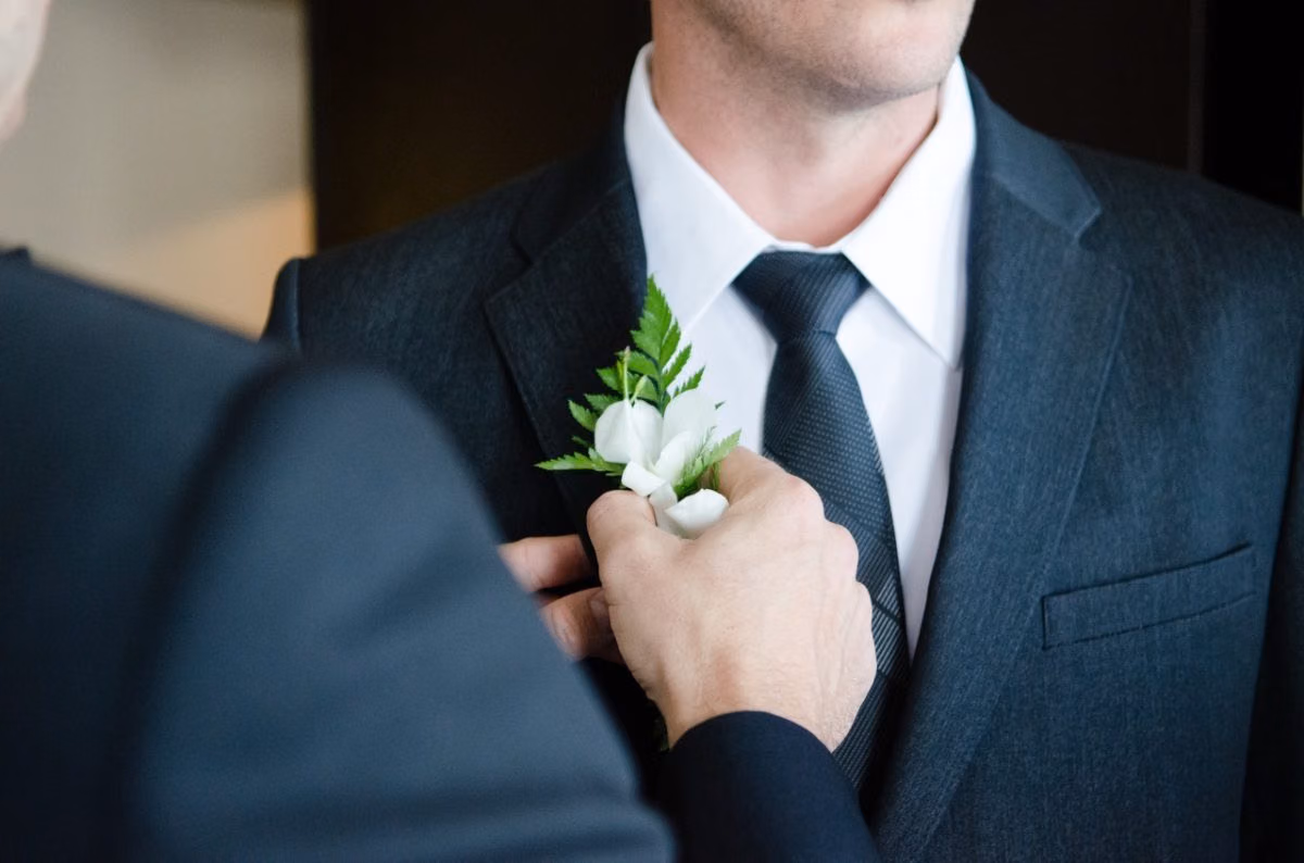 A groom pins a white flower boutonniere onto his partner's dark suit lapel