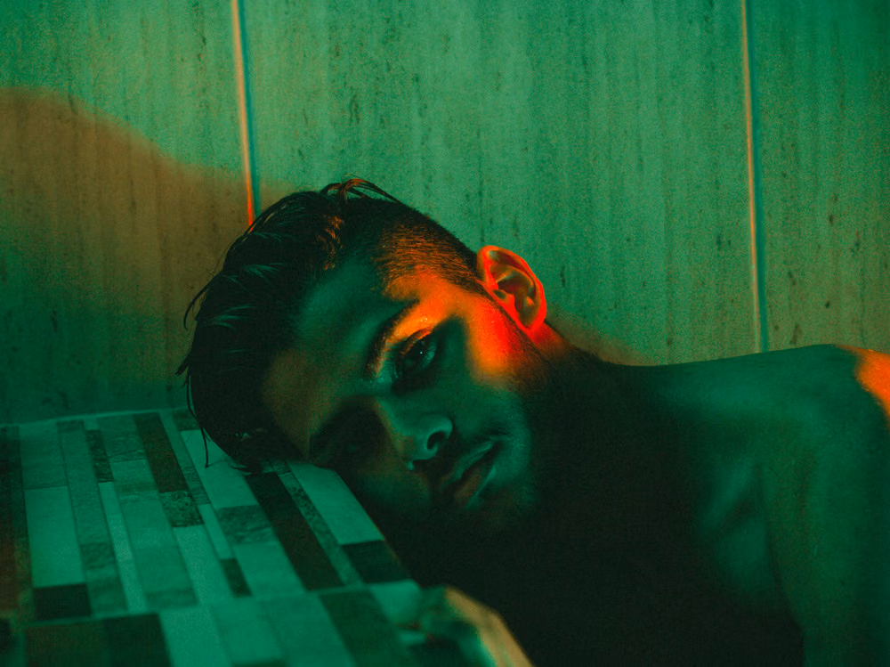 Shirtless man resting his head on a tiled ledge, lit in moody green and red light