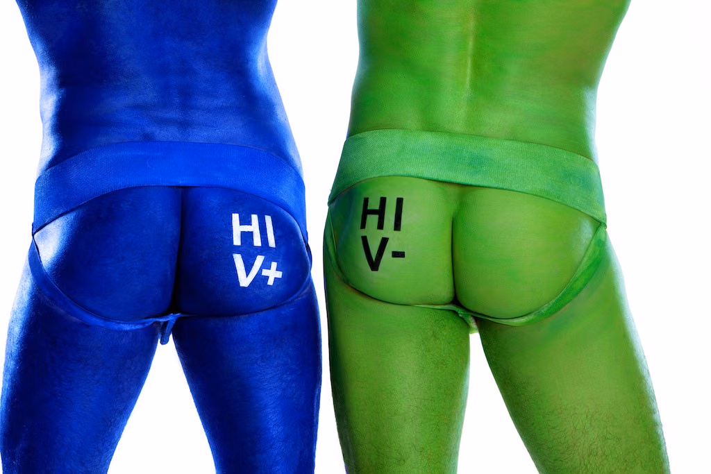 Two men in body paint and jockstraps seen from behind, one blue marked HIV+ and one green marked HIV-