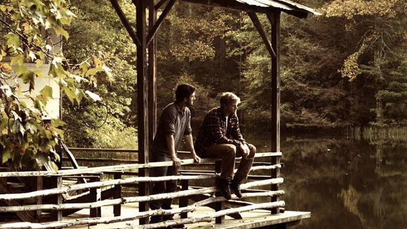 Two men on a rustic wooden dock overlooking a forest lake in a still from the gay film Before the Fall