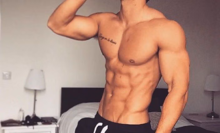 Shirtless muscular man with defined abs flexing his arm in a bedroom