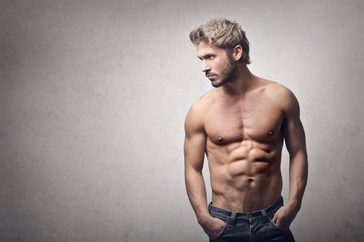 Shirtless fit man with tousled blond hair and hands in his jeans pockets against a gray wall