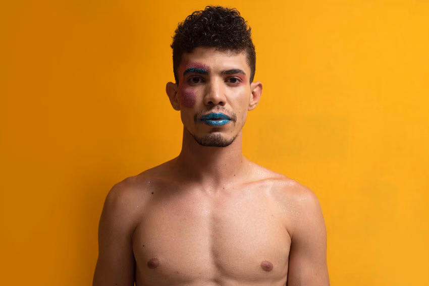 Shirtless young man with curly dark hair, pink and teal glitter makeup and teal glitter lips, against a bright yellow wall