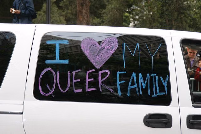 Hand-lettered message on a white car window reading "I love my queer family," with a purple heart standing in for "love"
