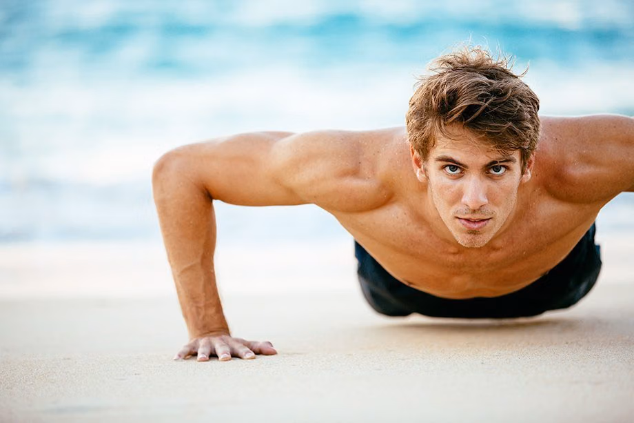 Shirtless man doing push-ups on the sand at the beach
