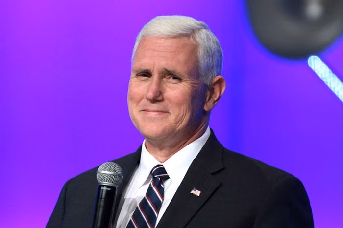 Mike Pence smiling on stage in a dark suit and striped tie, a microphone in front of him, against a purple backdrop
