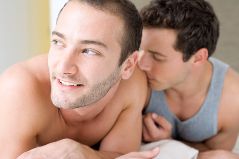 A smiling shirtless man looks away as another man nuzzles his shoulder
