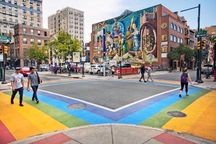 Rainbow crosswalks at 13th and Locust Streets in Philadelphia's Gayborhood, with pedestrians crossing below a large mural