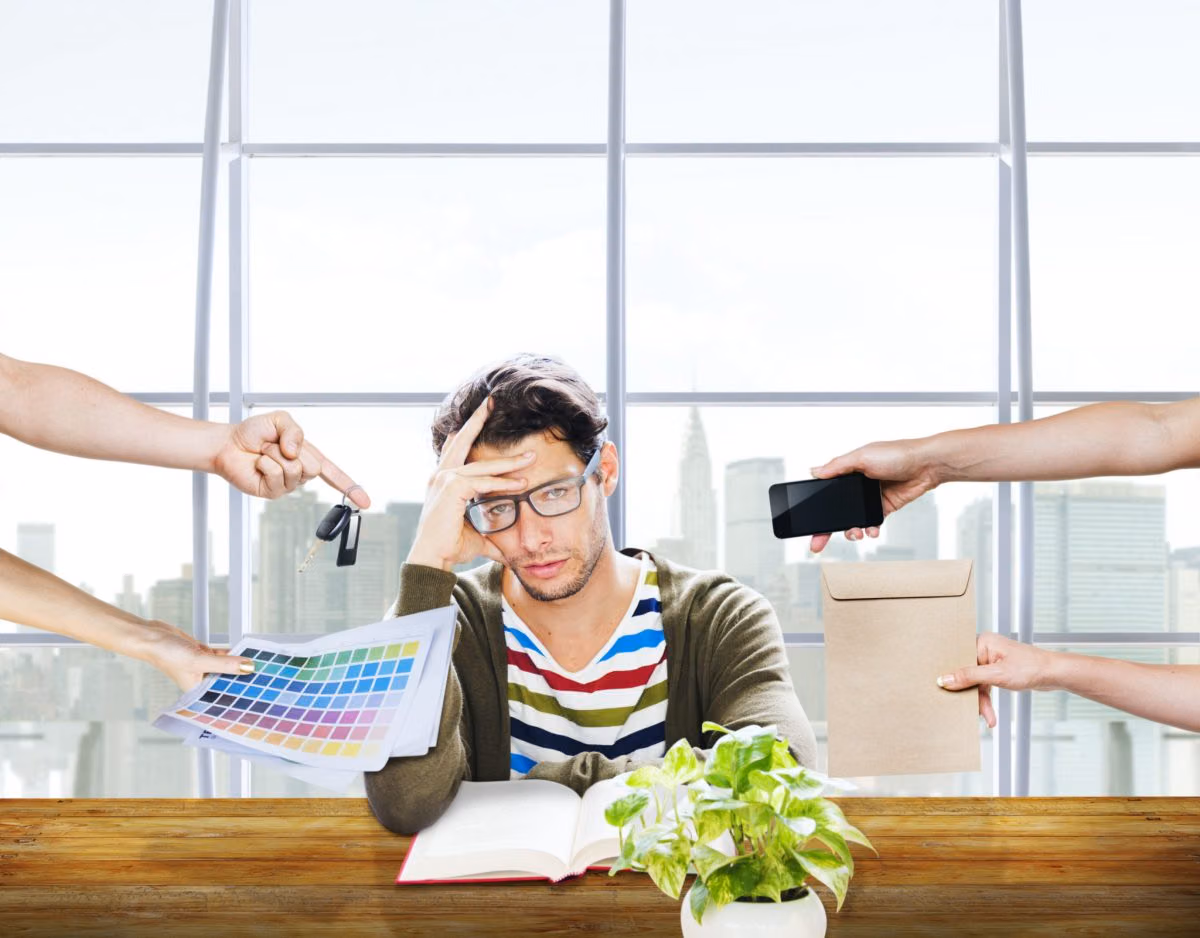 Stressed man at a desk holding his head as hands push keys, a phone, color swatches and an envelope at him