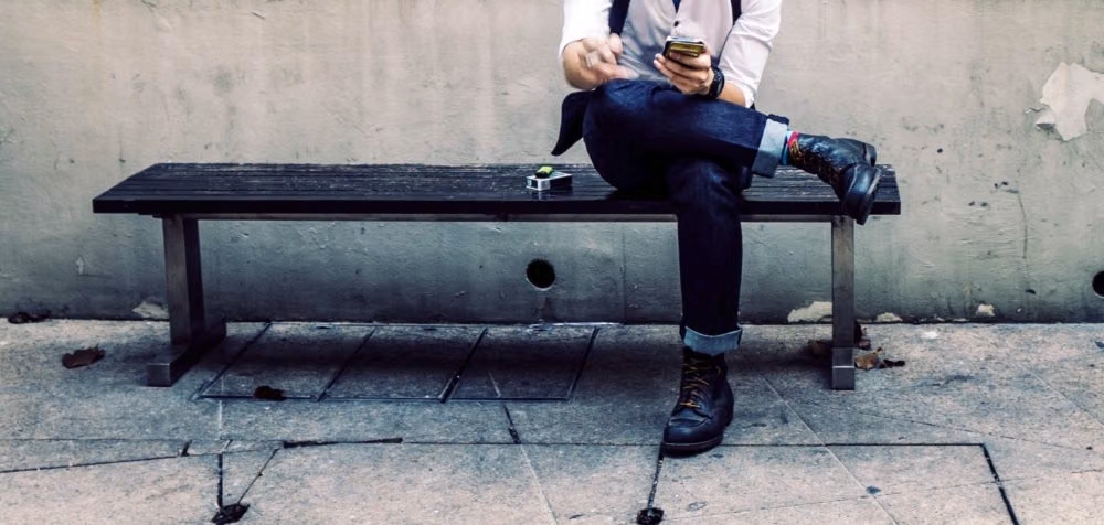 Man on a bench scrolling on his phone, face out of frame, against a bare concrete wall