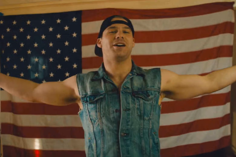 Young man in a backward cap and sleeveless denim shirt, arms spread before an American flag, in MTV's American Male