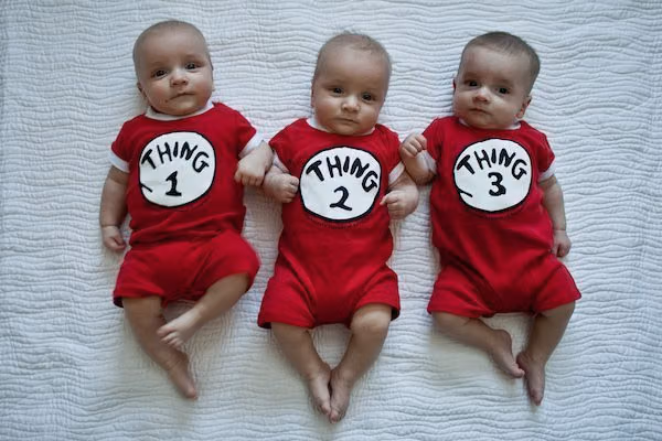 Three babies lying side by side on a white quilt in matching red Thing 1, Thing 2 and Thing 3 onesies