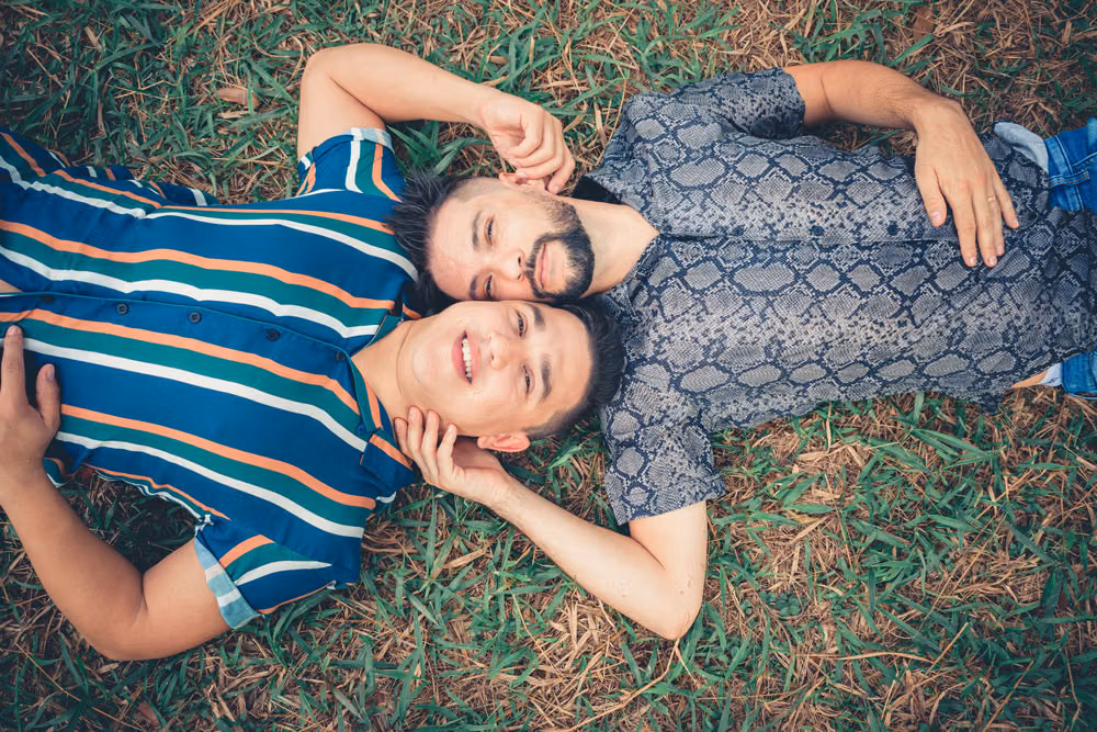 Two smiling men lying head to head on the grass, one in a striped shirt and the other in a snakeskin-print shirt