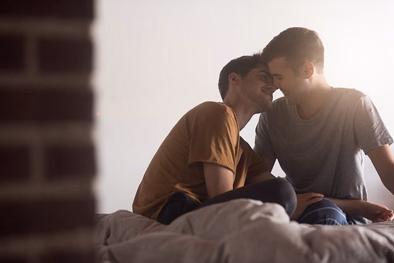 Gay couple in T-shirts sitting on a bed with foreheads touching, smiling