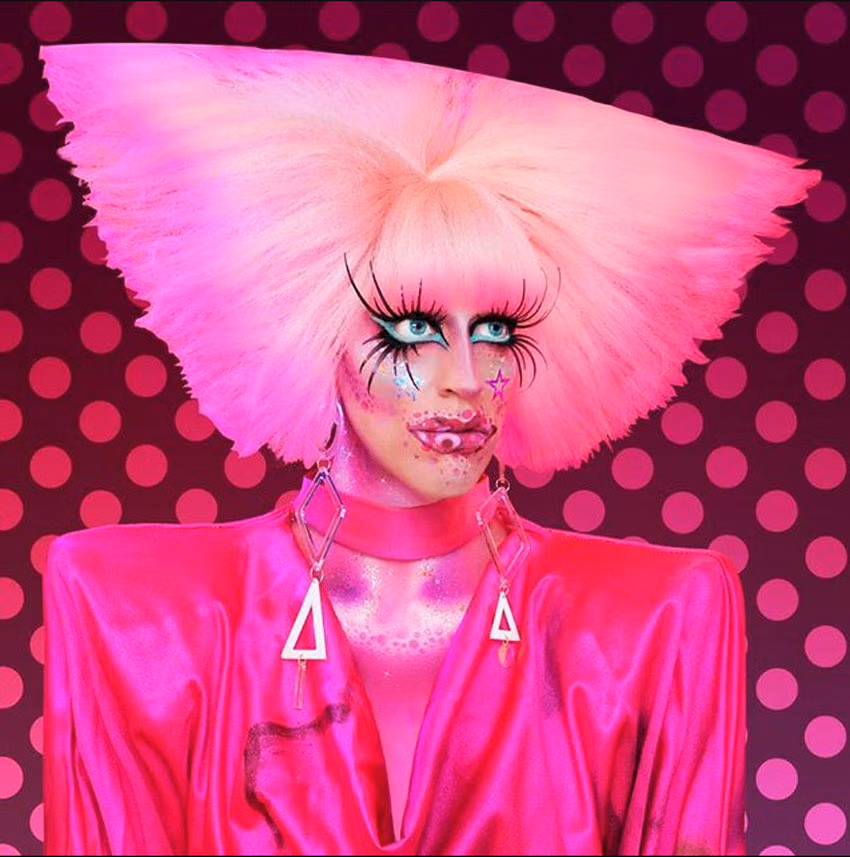 Acid Betty in a towering pink wig, alien-inspired makeup and a hot pink satin top against a pink polka-dot backdrop