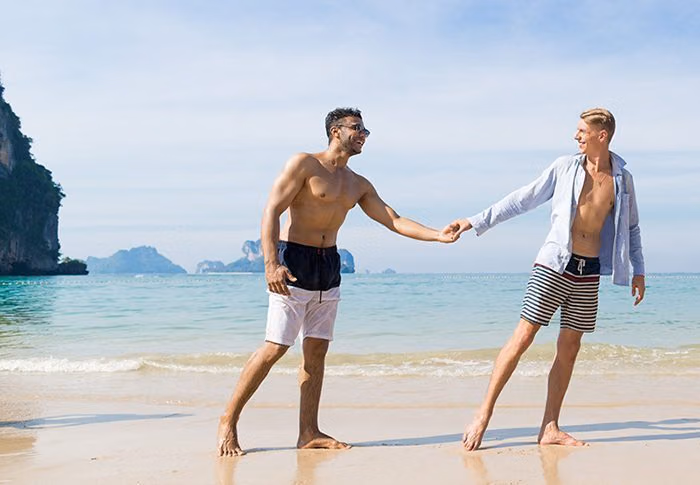 Gay couple holding hands on a tropical beach with limestone cliffs and turquoise water