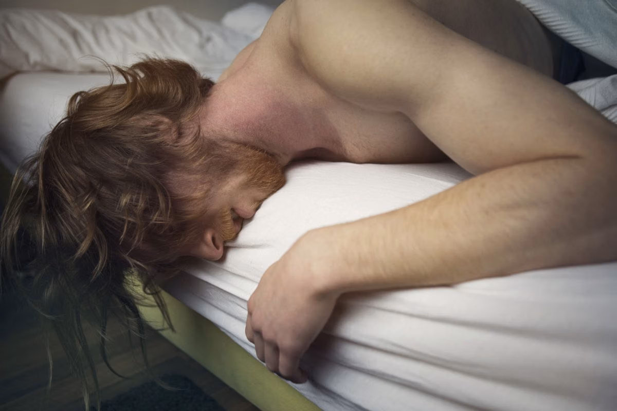 Shirtless man lies face down across the edge of a bed, his head hanging over the side