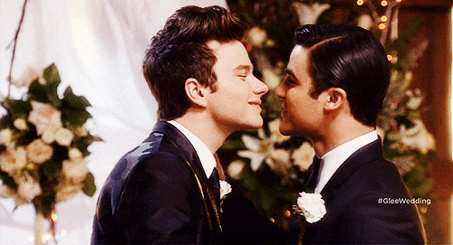 Two grooms in tuxedos smile and lean in for a kiss at a Glee wedding