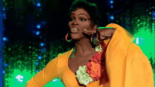 Coco lip-syncing in a yellow dress with a floral corsage, tapping her cheek, then Michelle Visage reacting