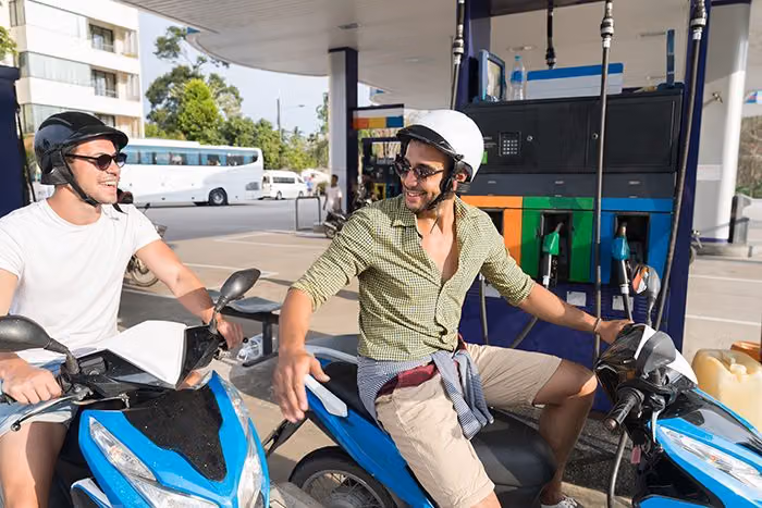 Two men in helmets laughing on blue scooters at a gas station during a trip