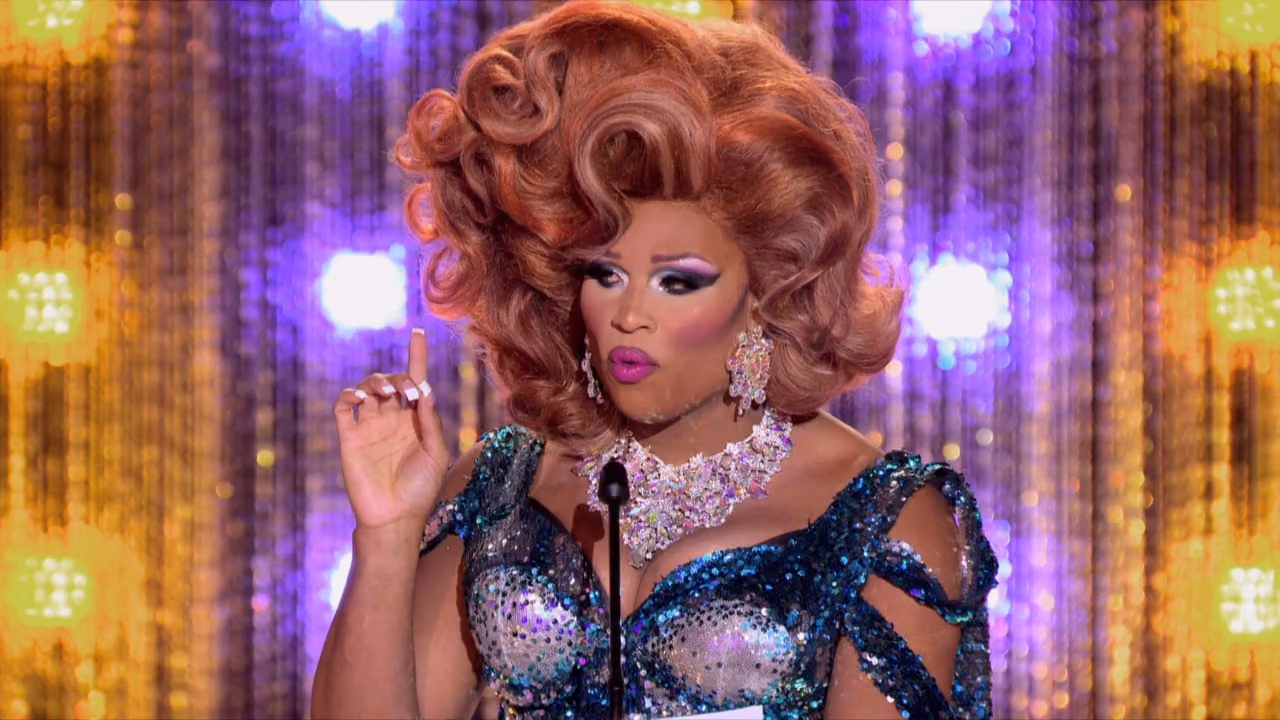 Drag queen in a teal sequined gown and voluminous auburn wig delivering a joke at the roast podium