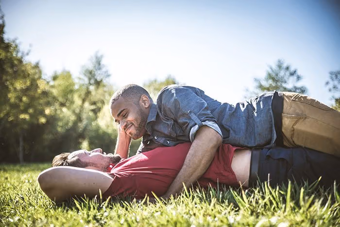 Two men lying in the grass in a sunny park, one resting on the other and smiling