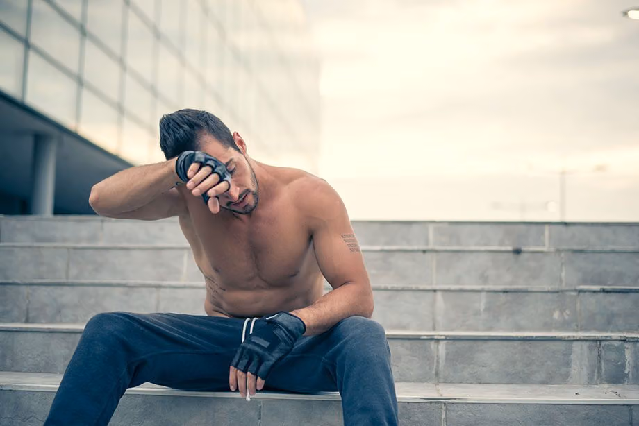 Tired shirtless man in workout gloves resting on outdoor steps, wiping his brow