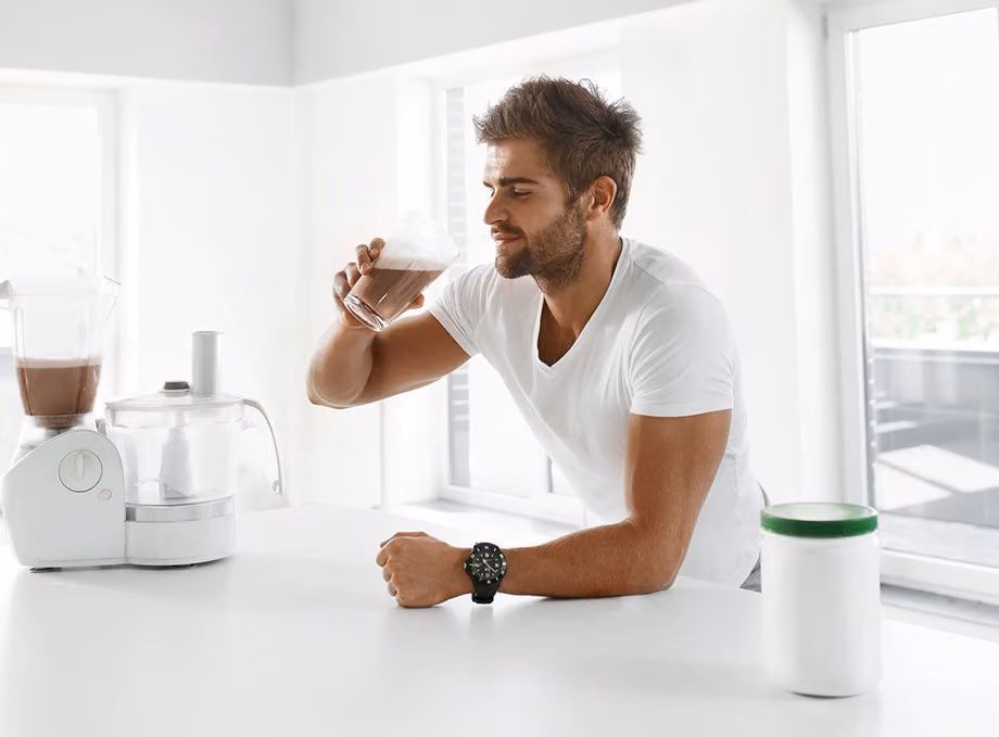 Man drinking a chocolate protein shake at a kitchen counter beside a blender