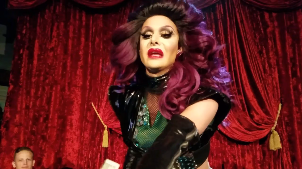 Trinity Taylor in a purple wig and black vinyl look, performing in front of a red velvet curtain