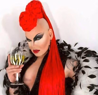 Nina Flowers with sharp black winged eye makeup and a red sculpted wig, holding a glass of white wine