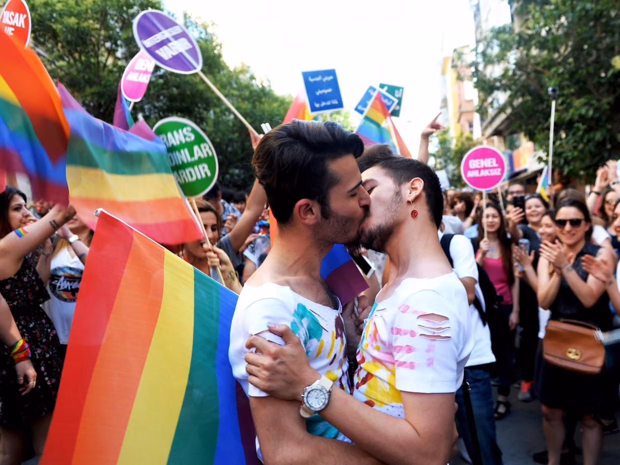 Two men kiss among rainbow flags and protest signs at Istanbul Pride as onlookers cheer