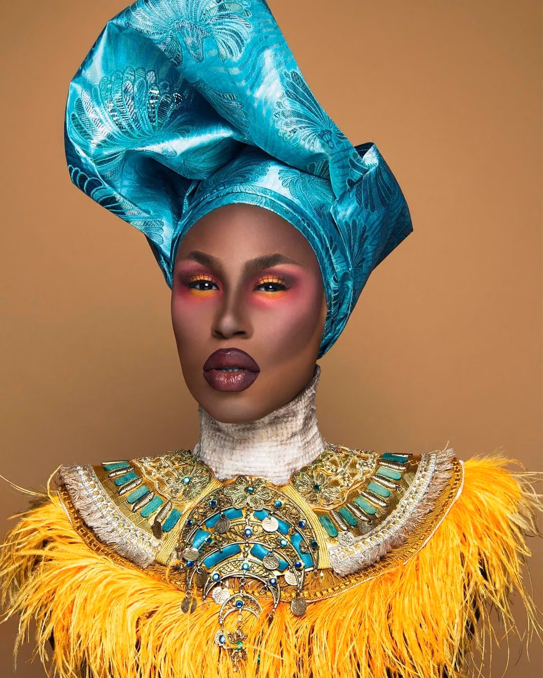 Shea Coulée in a teal head wrap, jeweled gold collar and yellow feathers