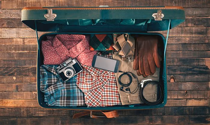 Open vintage suitcase packed with plaid shirts, a camera, phone, earbuds, leather gloves and a belt