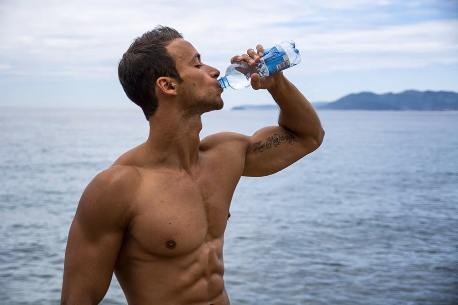 Shirtless man drinking bottled water by the sea, for the tip on staying hydrated