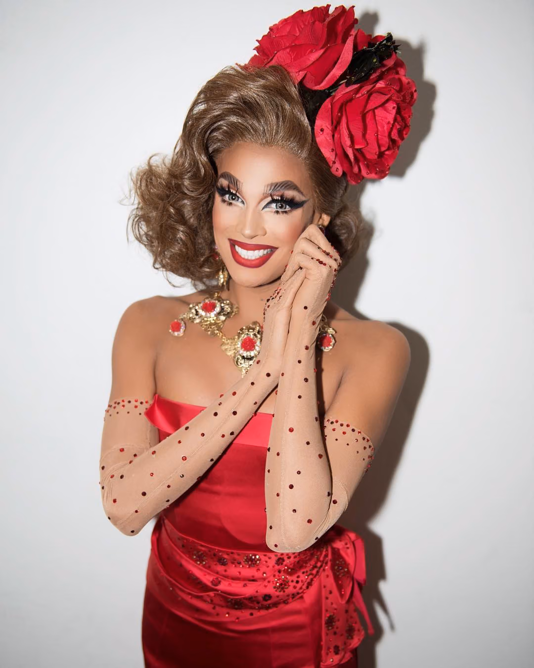 Smiling drag queen in a red strapless gown with a giant red rose headpiece and jeweled gloves