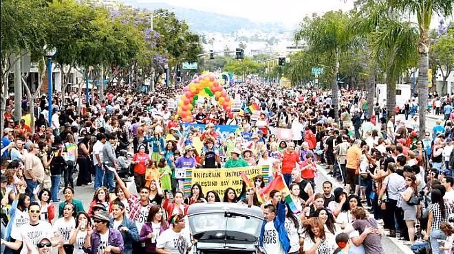 Crowds line a palm-lined boulevard as marchers carry banners and rainbow balloons at LA Pride