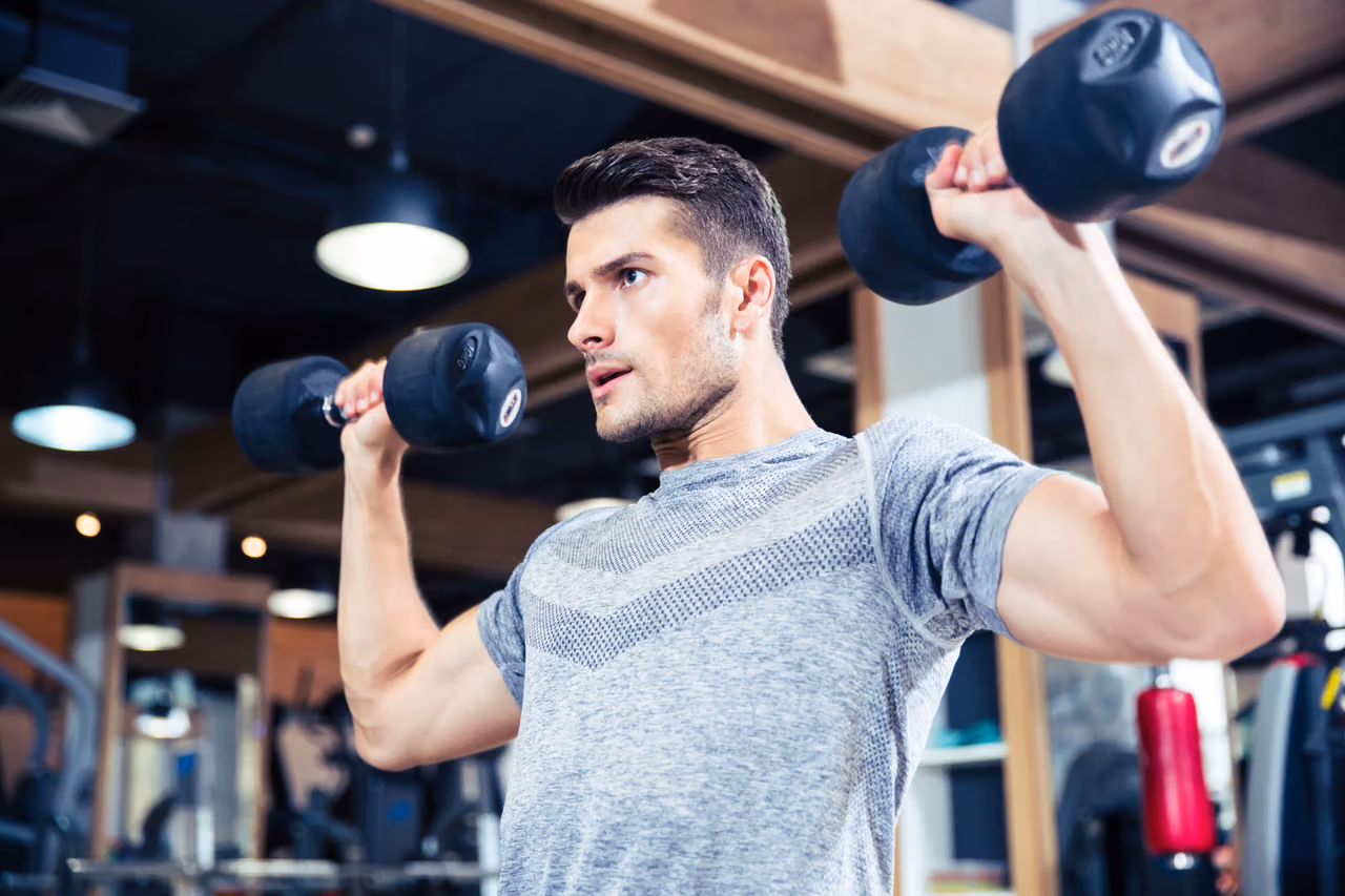 Man in a grey T-shirt presses two dumbbells overhead in a gym