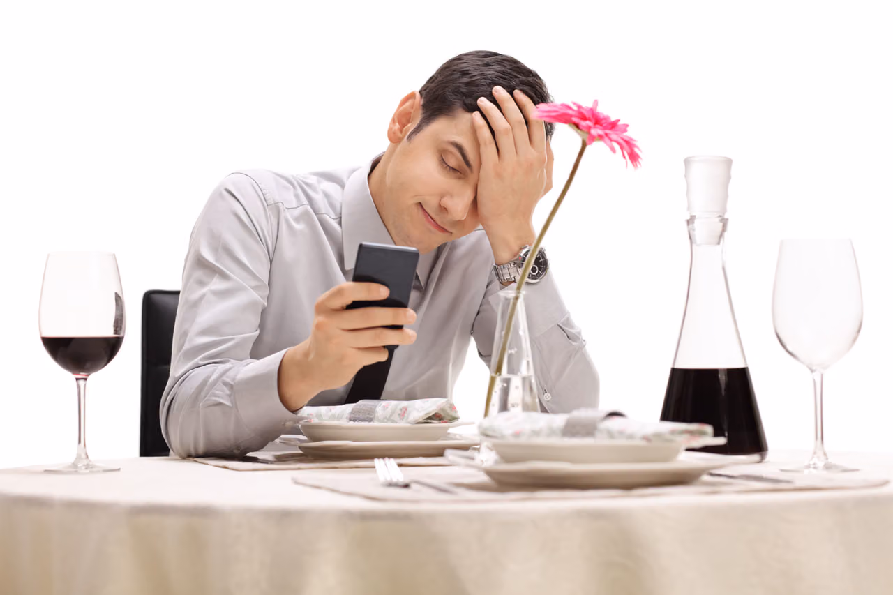 Man at a restaurant table set for two holds his phone and presses a hand to his forehead
