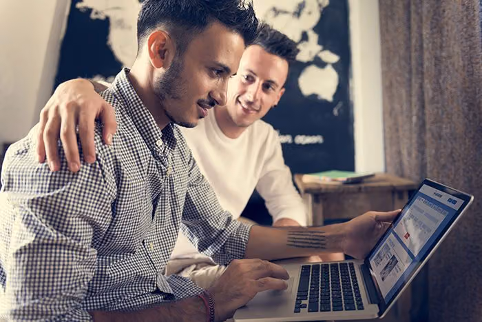 Two men smiling at a laptop as they plan a trip together, a world map on the wall behind them