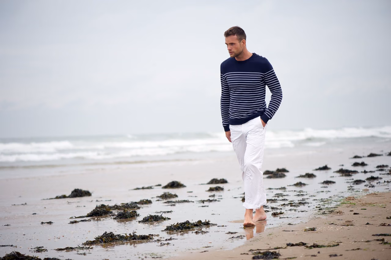 Man in a striped sweater walks barefoot along a beach, deep in thought