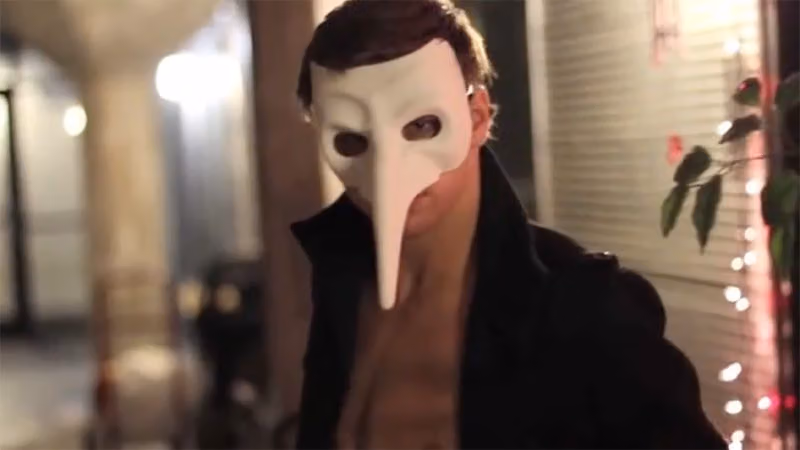 Man in a white long-nosed mask and open black coat on a street at night, from the Black Spark films