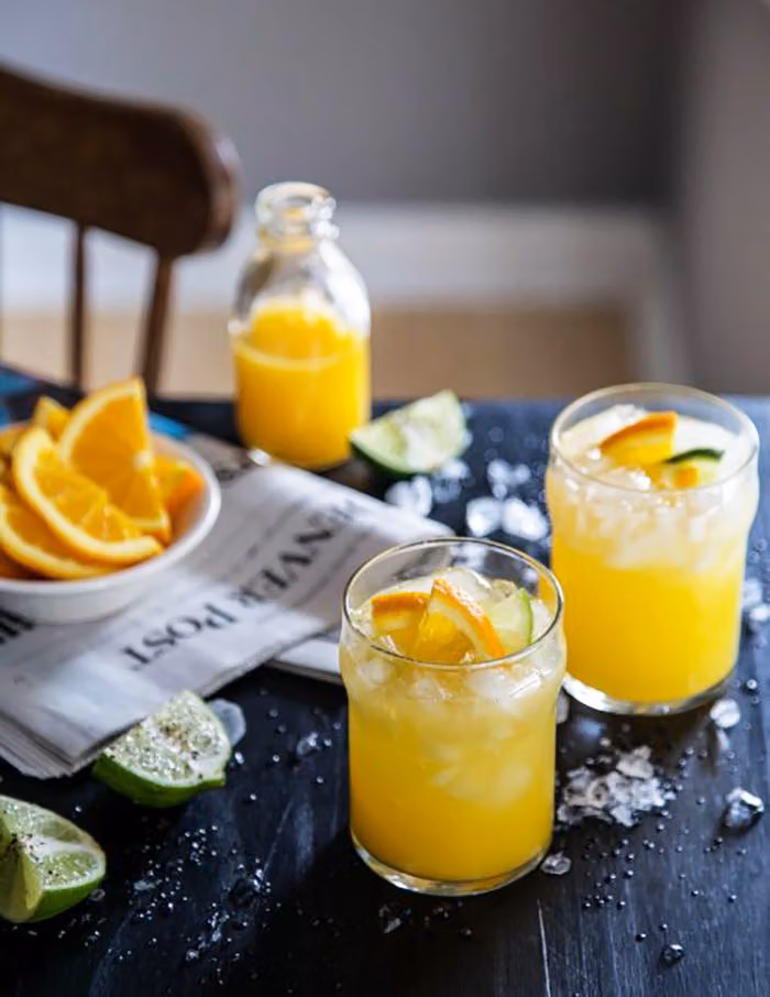 Two orange juice cocktails over ice with orange slices, beside a bottle of juice and lime wedges