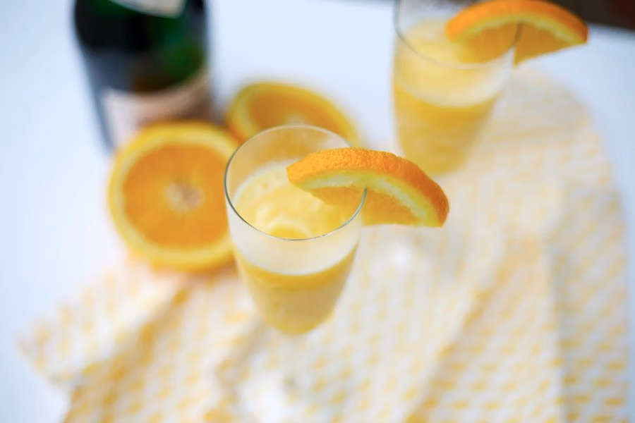Mimosas in champagne flutes garnished with orange slices, a bottle of sparkling wine behind them