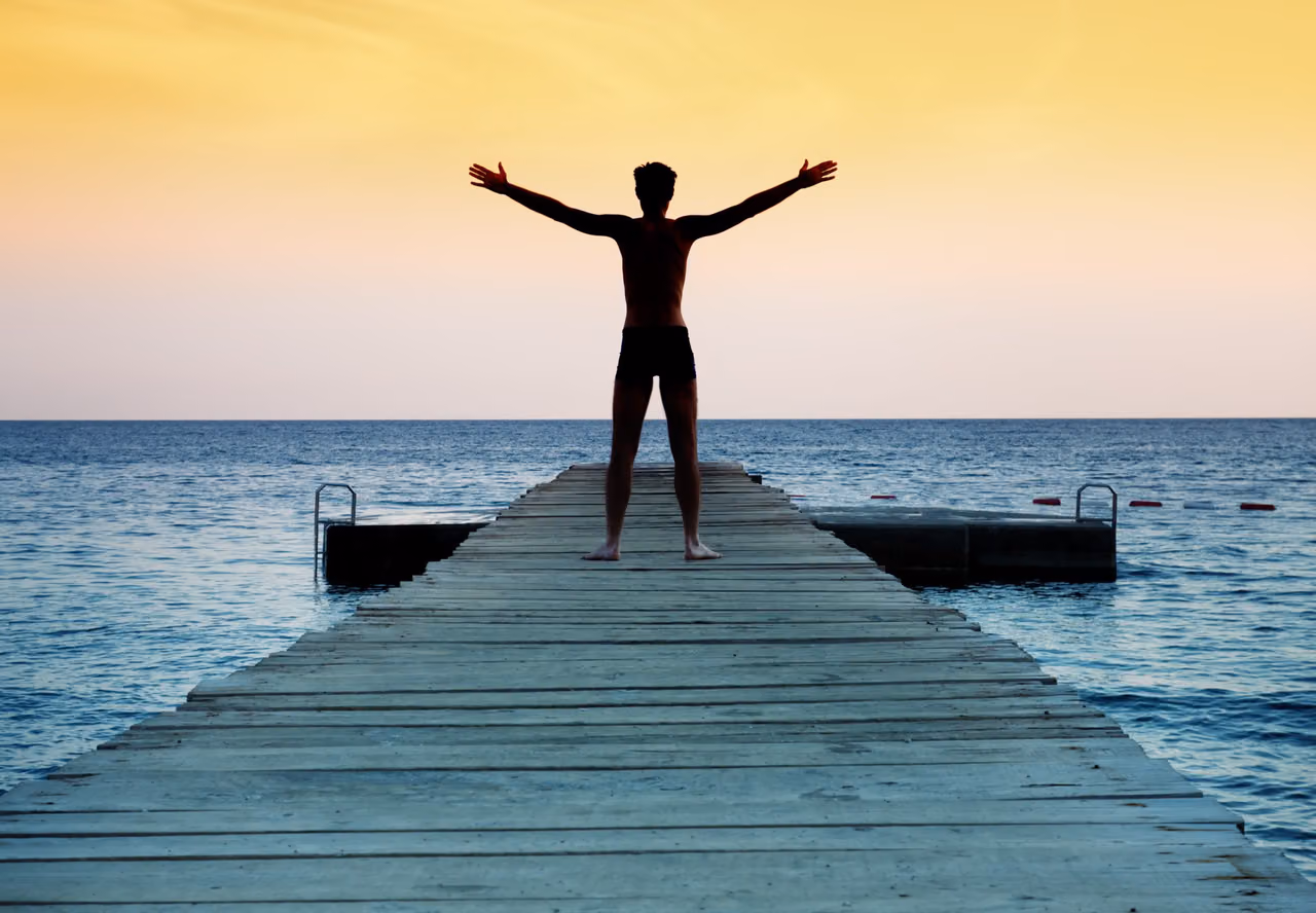 Man in swim trunks stands at the end of a pier with arms outstretched toward a sunset sea